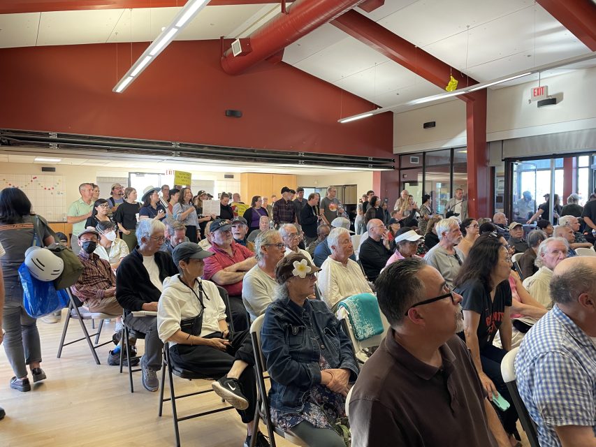 A large group of people, mostly older adults, sit and stand in a community center room during a public meeting or event.