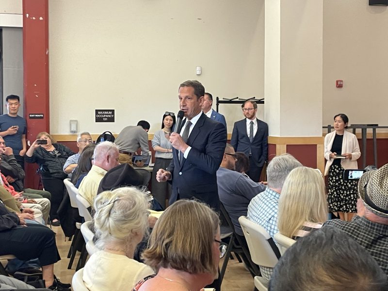 A man in a suit speaks into a microphone to a seated audience at an indoor event, while others stand and observe in the background.