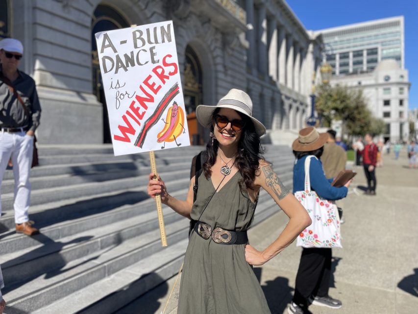 A woman wearing sunglasses and a hat stands outside holding a protest sign that reads "A-bun dance is for wieners" with a drawing of hot dogs.