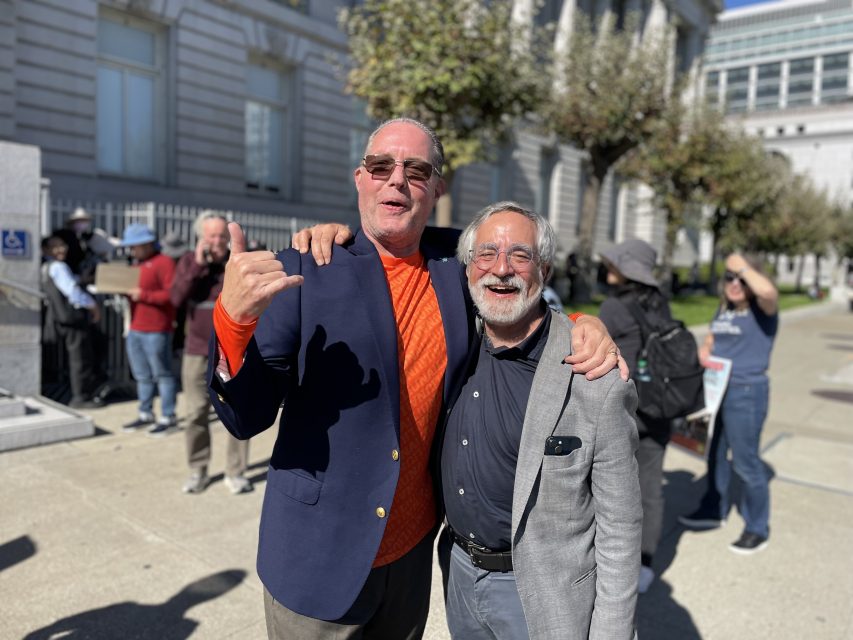 Two older men stand arm in arm and smiling outdoors in a city setting, with other people and buildings visible in the background.