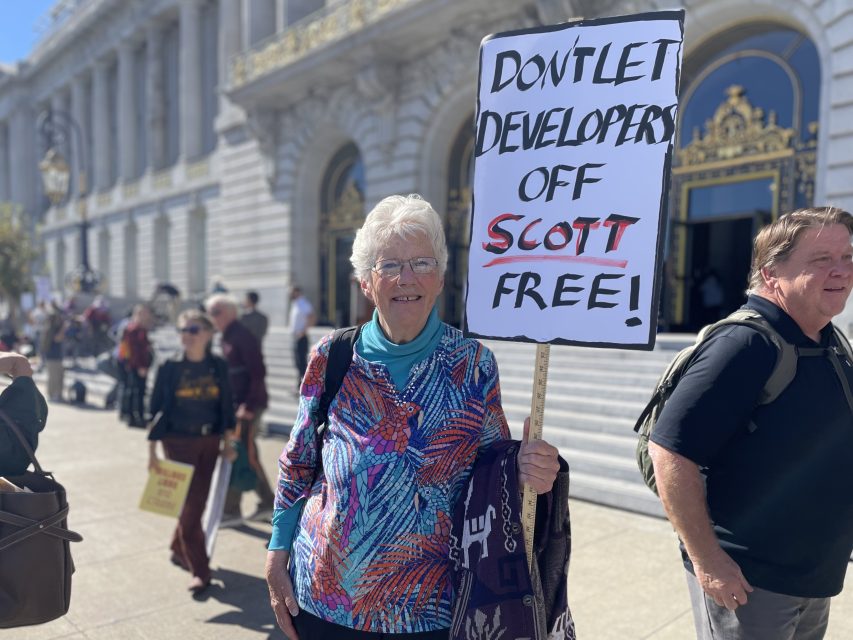 An older woman stands outside holding a sign that reads, "Don't let developers off Scott free!" Other people are visible in the background.