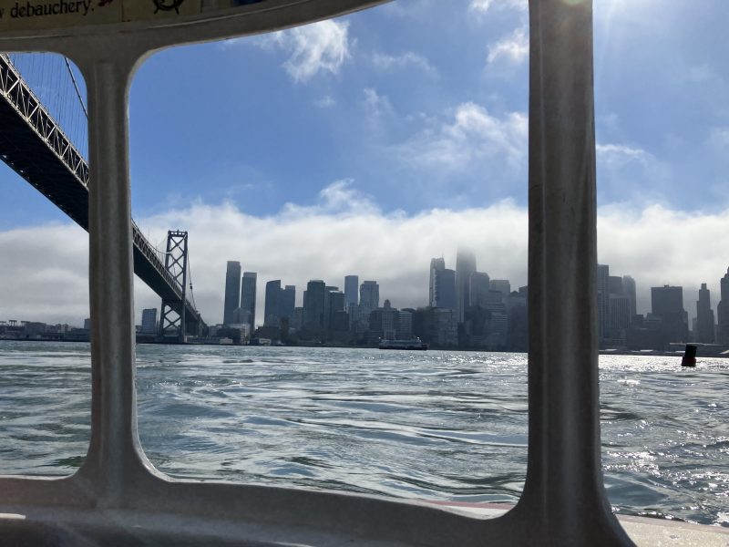 View of the San Francisco skyline and Bay Bridge seen through the window of a boat on a partly cloudy day.