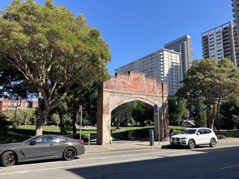 A brick archway stands on a sidewalk between two parked cars, with trees and modern high-rise buildings in the background under a clear blue sky.