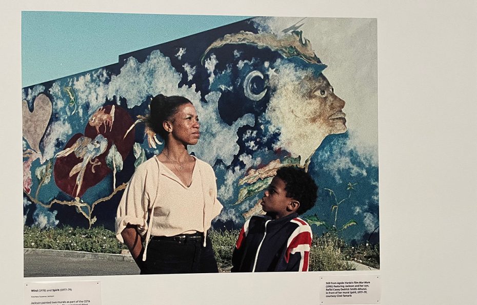 A woman and a child stand in front of a colorful mural featuring abstract human faces and shapes painted on an outdoor wall.