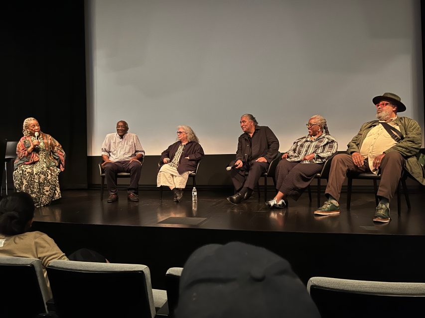 Six people sit on stage in front of a projection screen, engaged in a panel discussion. Microphones and a water bottle are visible; audience members are in the foreground.