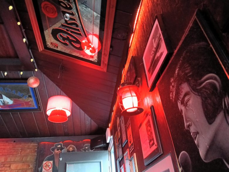 Wall decorated with portraits, framed photos, and posters, illuminated by red lights and lamps, in a dimly lit room with a slanted wooden ceiling.