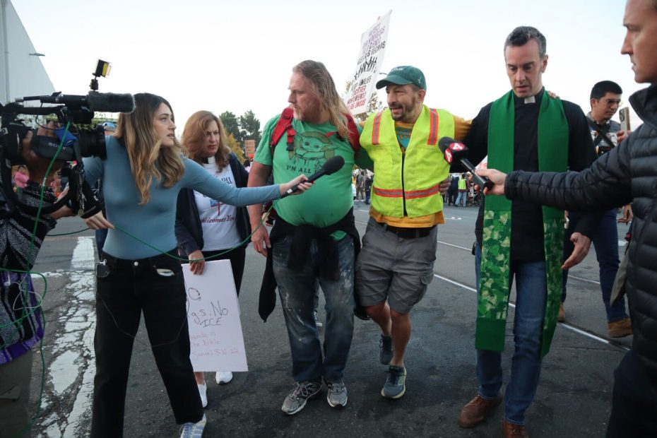 A group of people, some holding microphones and cameras, surround two men—one in a yellow vest and another in a green shirt—during a street protest focused on immigration issues.