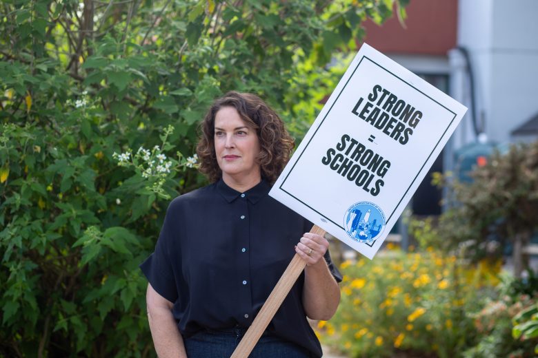 A person stands outdoors holding a sign that reads, "STRONG LEADERS = STRONG SCHOOLS," with plants and flowers visible in the background.