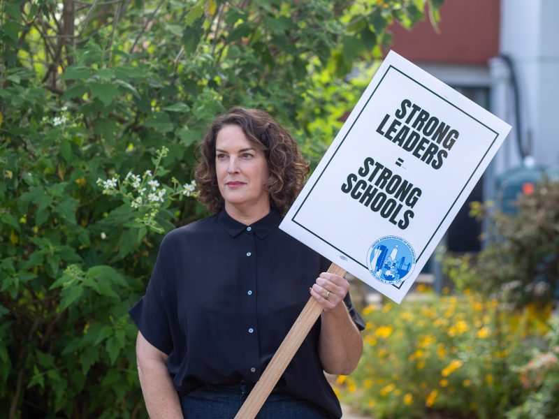 A person stands outdoors holding a sign that reads, "STRONG LEADERS = STRONG SCHOOLS," with plants and flowers visible in the background.