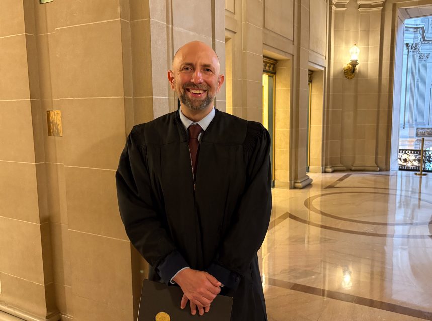 A man wearing a black judicial robe stands smiling in a grand, marble hallway with high ceilings and ornate lighting.