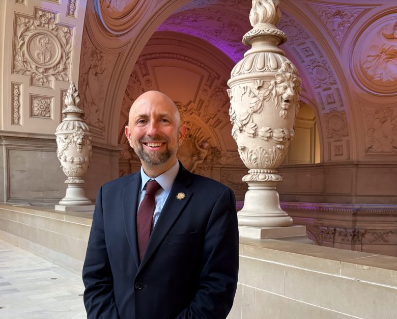 A smiling man in a suit and tie stands indoors in front of ornate architectural columns and detailed carvings, under purple and warm lighting.