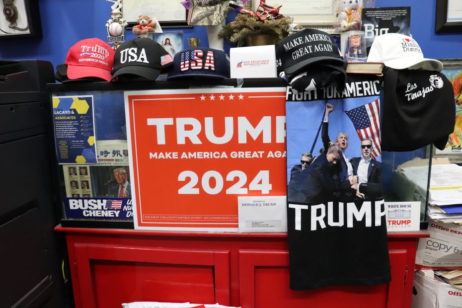 A display of Trump 2024 campaign signs, hats, and merchandise arranged on a red counter in an office setting.
