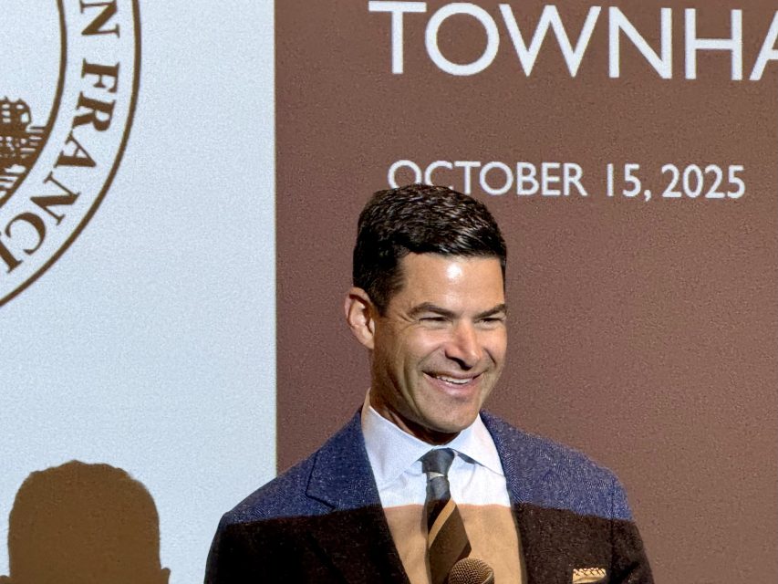 A man in a suit and tie stands in front of a projected screen displaying “TOWNHALL” and “October 15, 2025,” with part of a seal visible on the left.