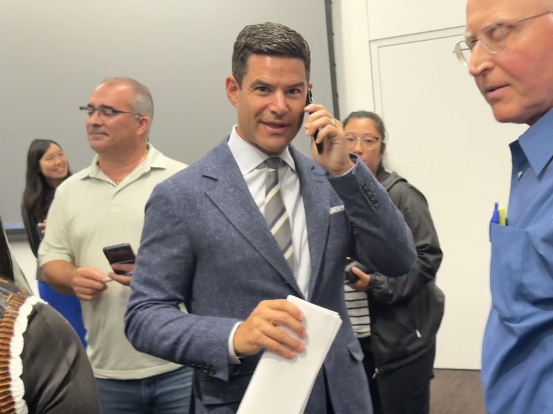 A man in a blue suit talks on the phone while holding papers in a crowded indoor setting with several people in the background.