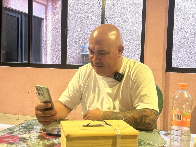 A man with a shaved head and tattoos looks at his phone while sitting at a table with a wooden box and a plastic bottle. He is wearing a white t-shirt and a lapel microphone.