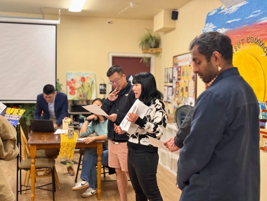 Four people stand and read from papers in a brightly lit room, with one person seated at a laptop in the background near a mural and bulletin board.
