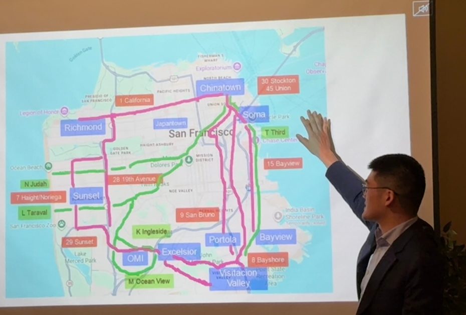 A man in a suit gestures toward a projected map of San Francisco neighborhoods highlighted with different colored routes and labels.