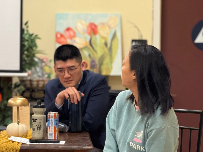 Two people sit at a table with drinks and papers; one faces the camera, the other looks at her. A painting of flowers hangs on the wall in the background.