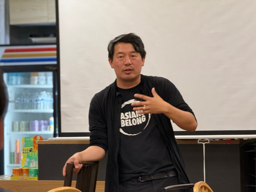 A person stands indoors in front of a white screen, gesturing with one hand and wearing a shirt that says "ASIANS BELONG.