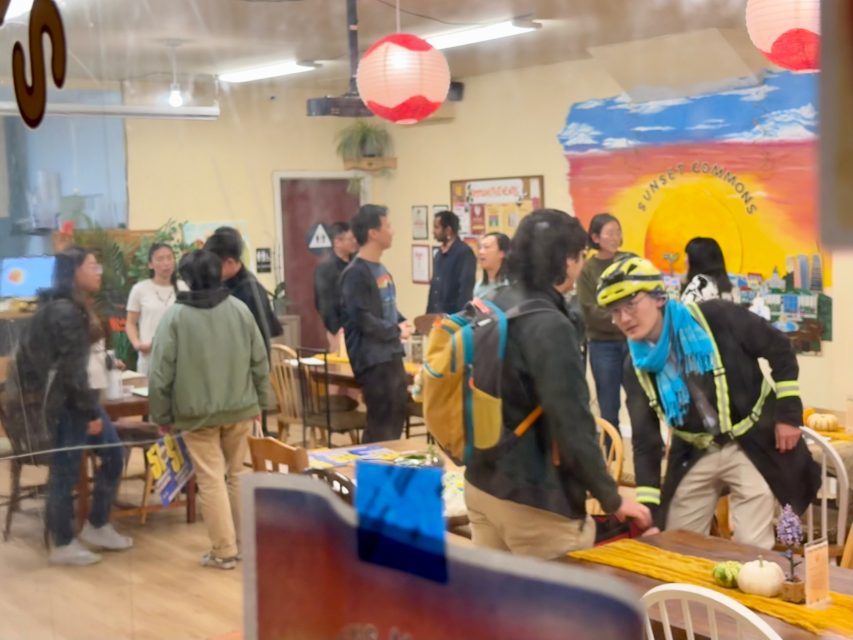 A group of people socializes in a casual indoor setting with tables and chairs; one person wears a bike helmet and jacket. A mural and decorations are visible on the wall.