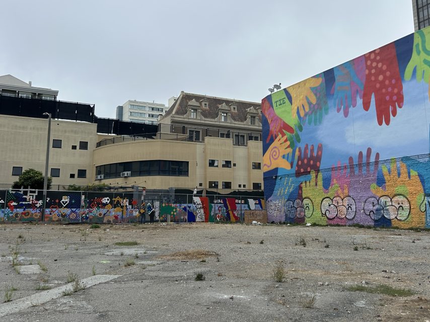A vacant lot with sparse weeds is bordered by colorful murals featuring abstract patterns, handprints, faces, and a soccer ball motif, with older buildings visible in the background on a cloudy day.