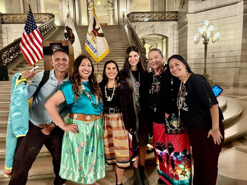Six people stand together and smile inside a grand building with ornate columns and flags, some wearing colorful skirts and traditional jewelry.