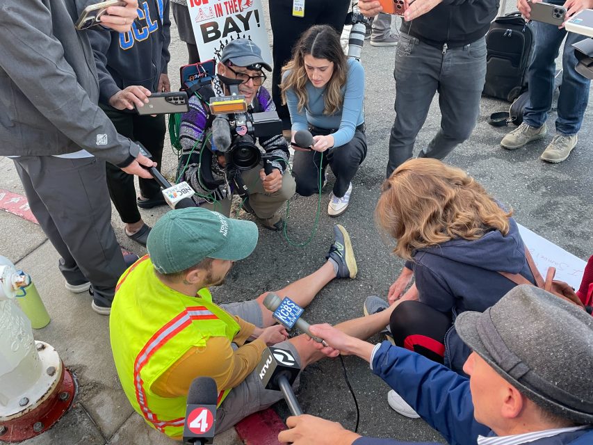 A man in a yellow safety vest sits on the ground surrounded by reporters with microphones, cameras, and notepads during a street interview about immigration.