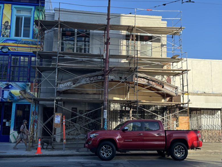 A red pickup truck is parked in front of a building under construction, covered in scaffolding. A person walks on the sidewalk nearby.