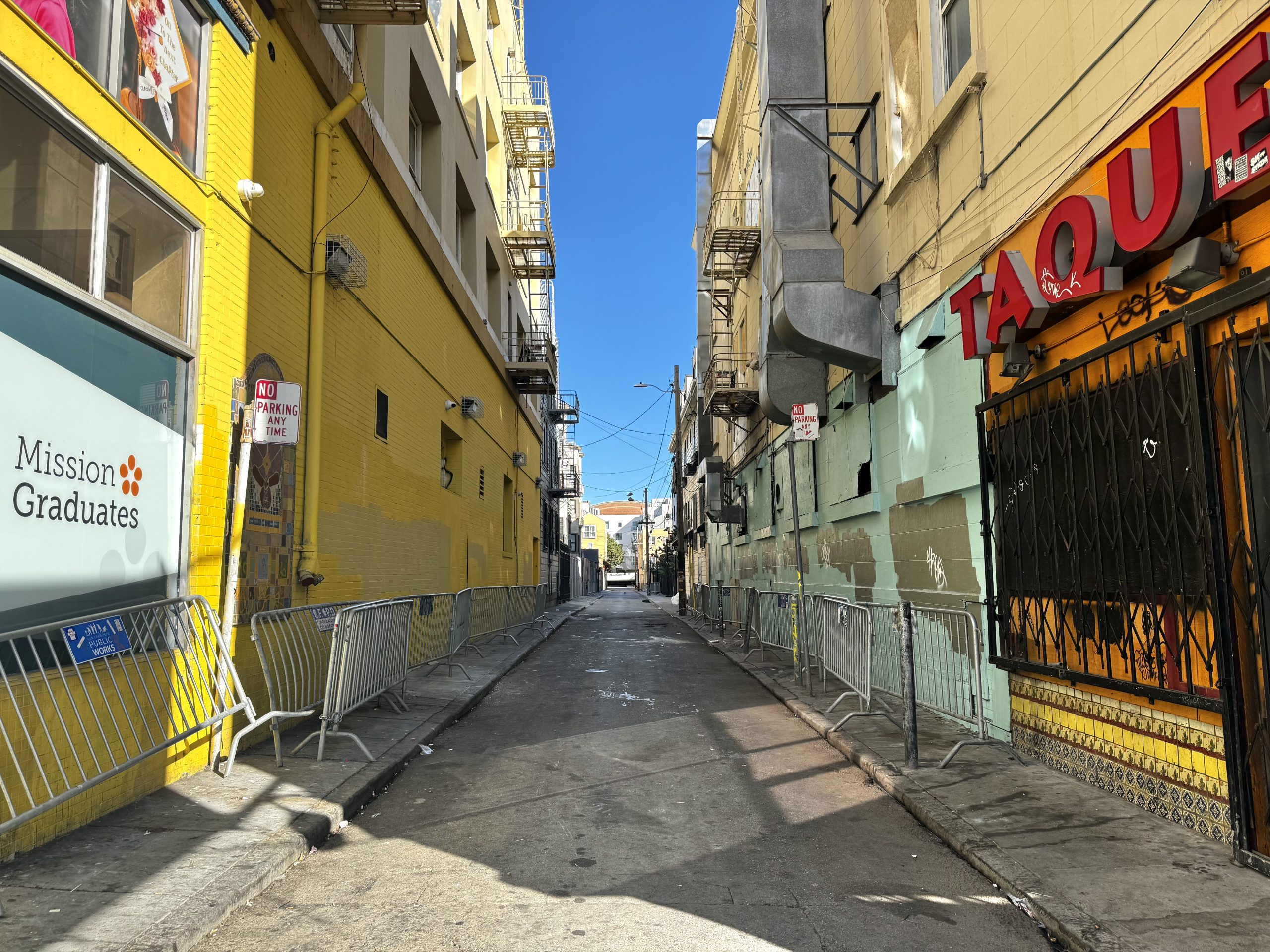A narrow city alley with metal barricades on both sides, yellow and beige buildings, and a “Mission Graduates” sign on the left. Bright sunlight and a clear blue sky overhead.