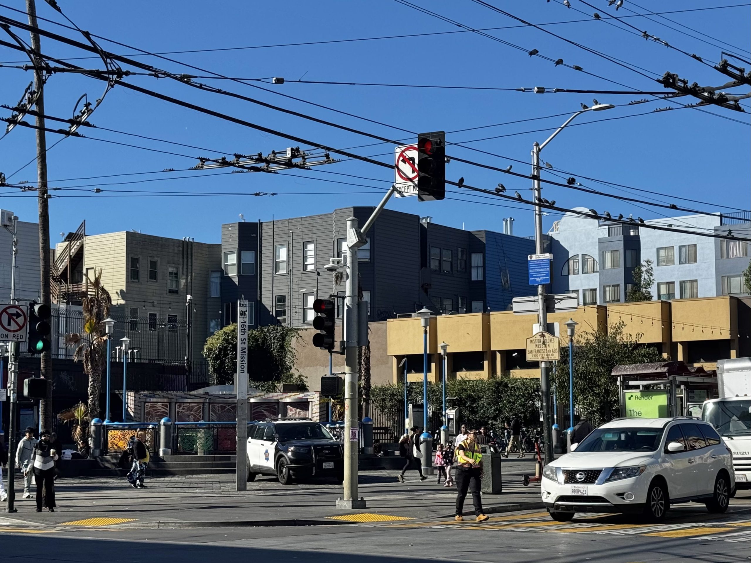 Urban street intersection with traffic signals, vehicles, pedestrians, and overhead power lines against a backdrop of multi-story buildings on a clear day.