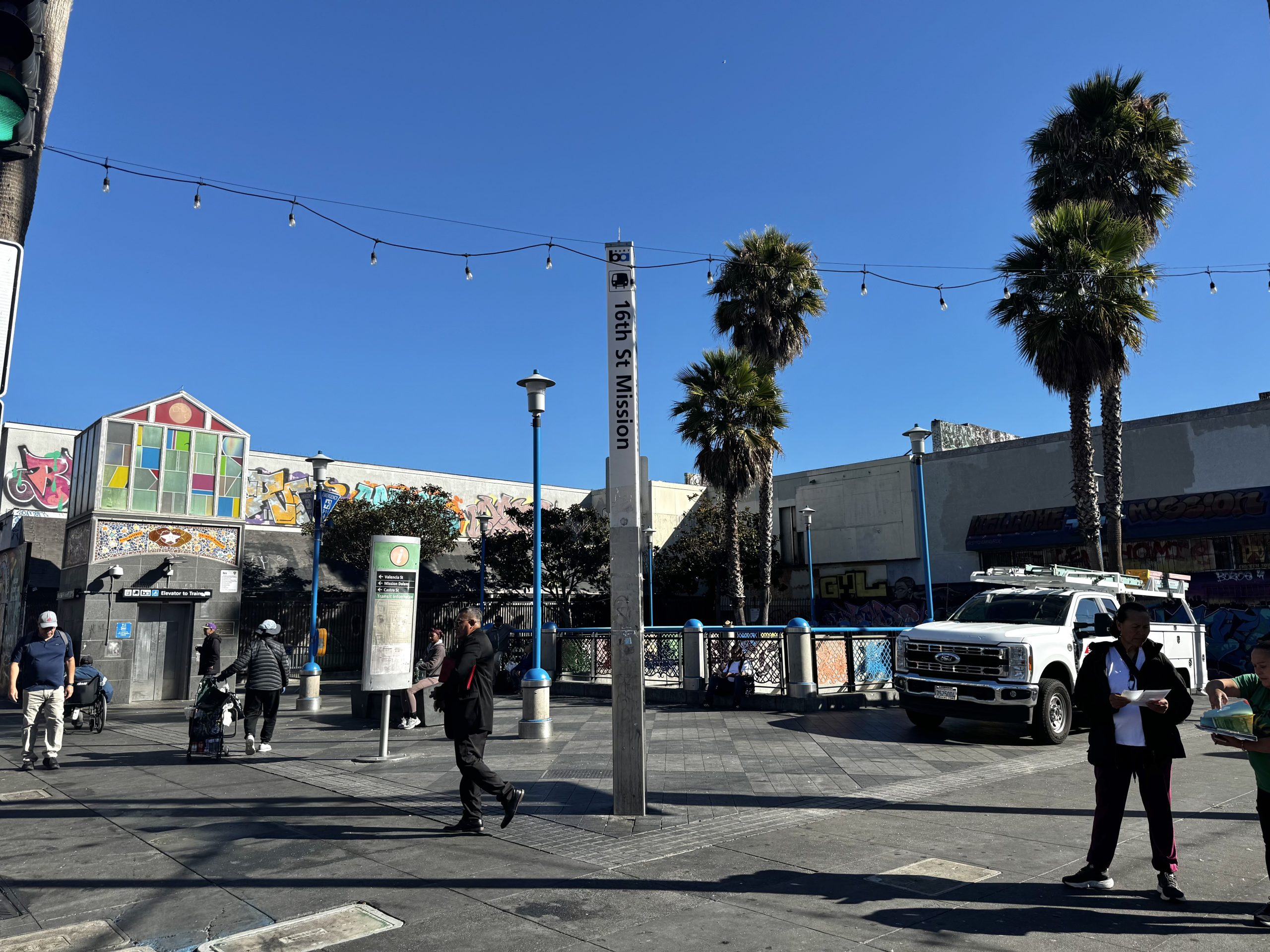 People walk near the 16th St Mission station sign in a sunny urban plaza with palm trees, graffiti, and a parked utility truck nearby.