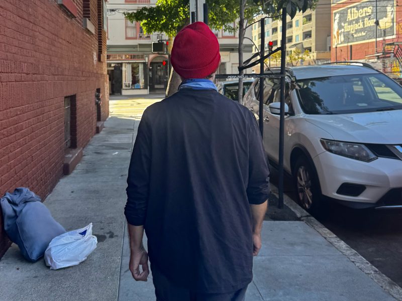 A person wearing a red beanie and black jacket walks down a city sidewalk next to parked cars and a brick building.