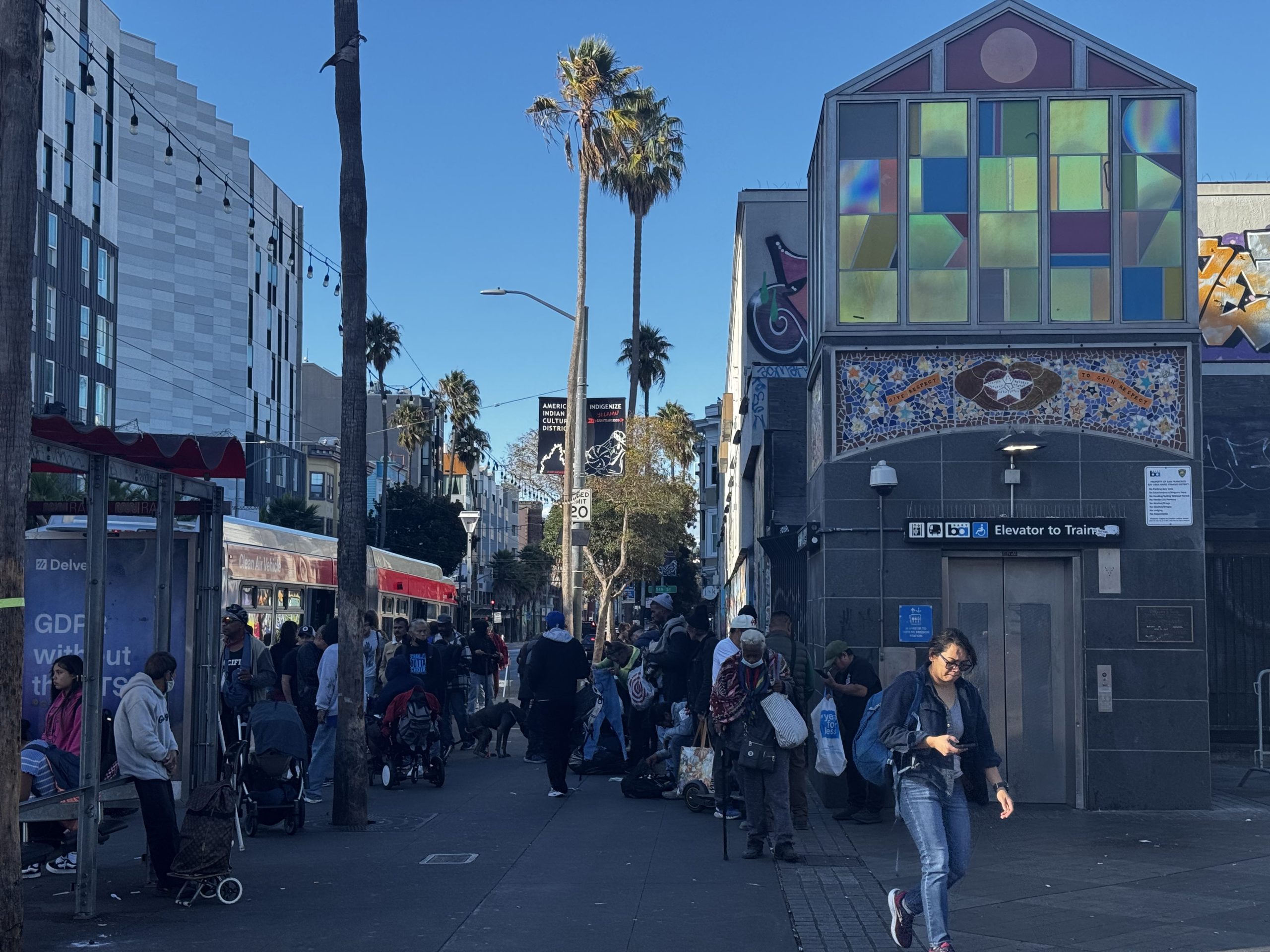 A busy urban sidewalk near a public transit entrance, with groups of people, palm trees, street art, and a bus stopped on the left.