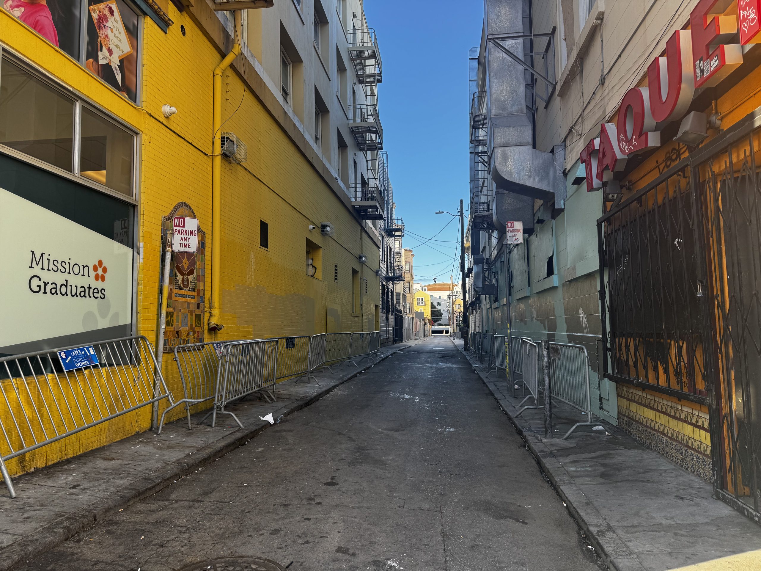 A narrow urban alleyway with metal barricades, yellow painted wall on the left, and various signs. The sky is clear and blue.