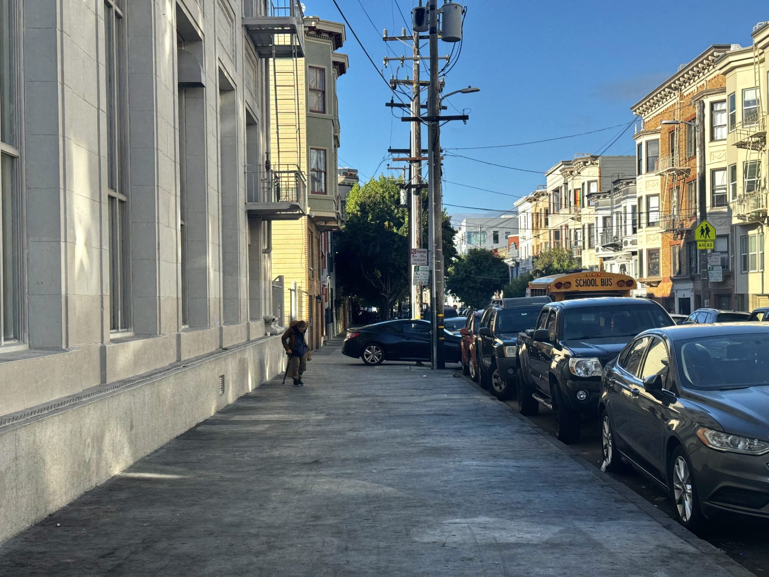 A city sidewalk with parked cars, a school bus, and a lone pedestrian. Multi-story buildings line both sides of the street under a clear blue sky.