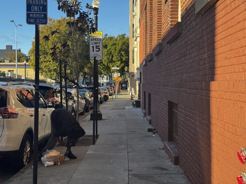 A person bends down near parked cars on a city sidewalk beside a brick building. Various street signs, including a speed limit sign, are visible.