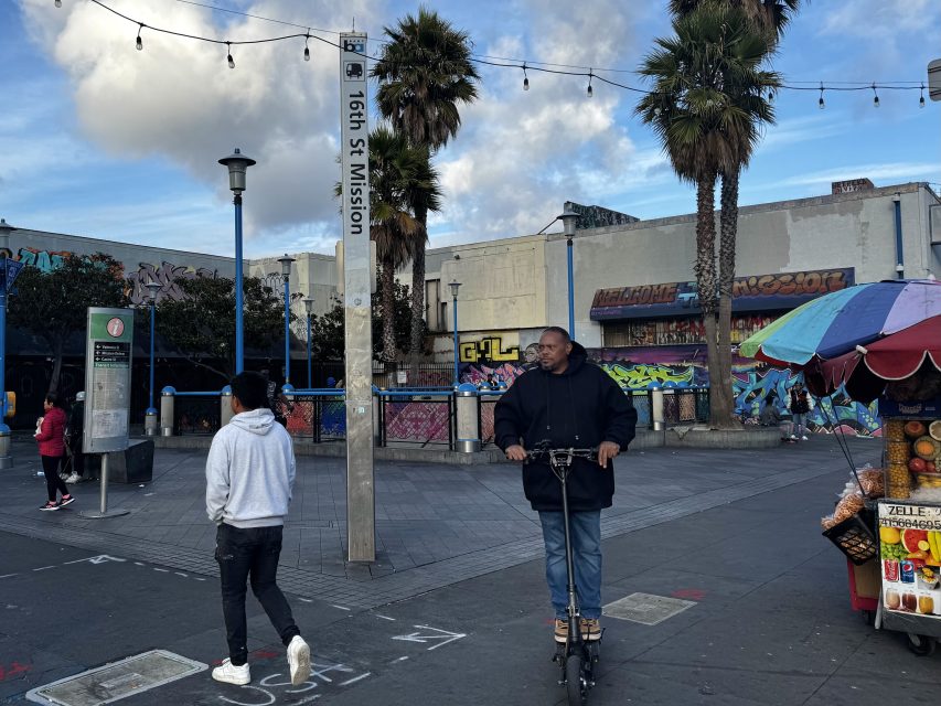 A man rides an electric scooter near the 16th St Mission sign while another person walks by; colorful murals and market stalls are in the background.