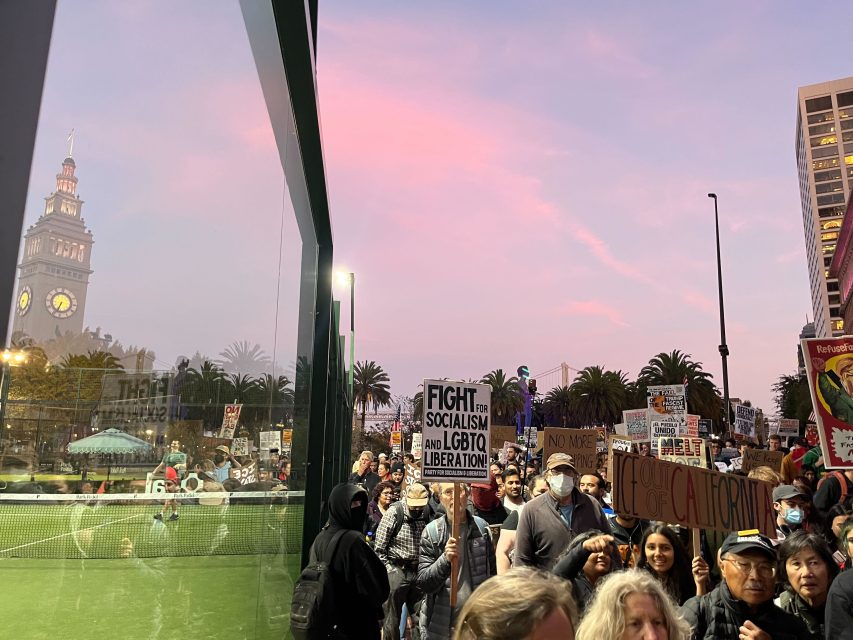 A crowd of protesters holding various signs, including one reading "Fight for Socialism and LGBTQ Liberation," stands near a glass wall in an urban area at dusk.