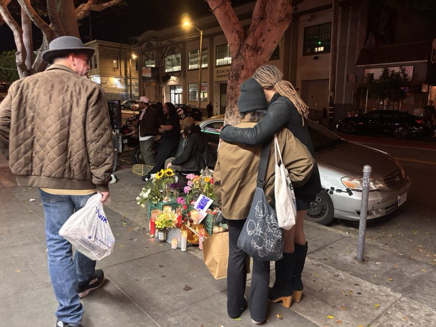 Two people hug beside a street memorial with flowers and candles at night, while others gather nearby; a man walks past carrying a plastic bag.