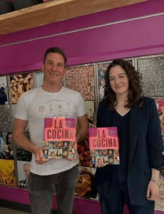 A man and a woman stand indoors, each holding a copy of a book titled "La Cocina" in front of a wall with food-themed photos.