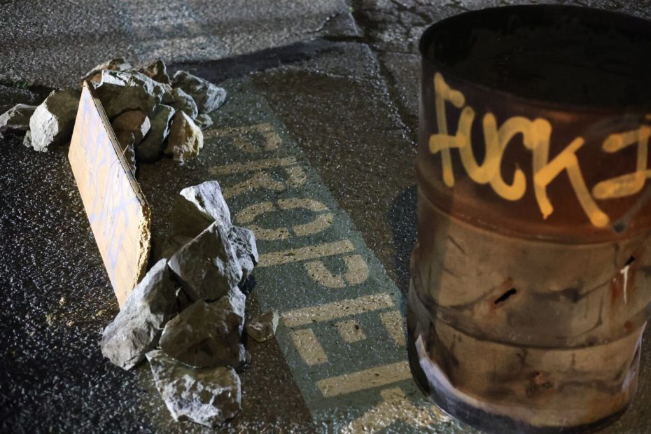 A metal barrel with graffiti stands on asphalt next to rocks and a broken property sign.