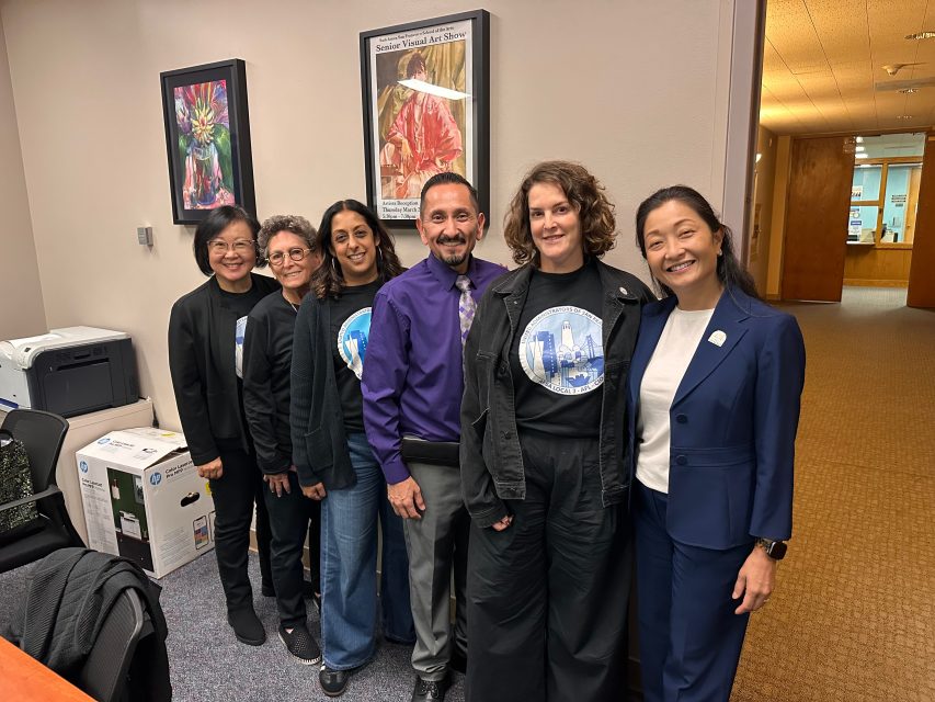 Six people stand side by side in an office, some wearing matching cityscape shirts, with framed artwork on the wall and office equipment in the background.