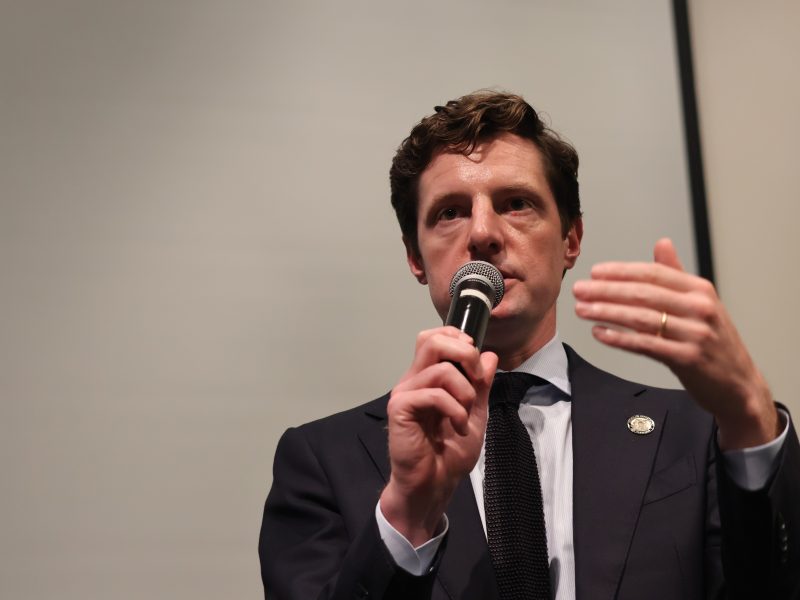 A man in a suit speaks into a microphone while gesturing with his hand in front of a plain background.