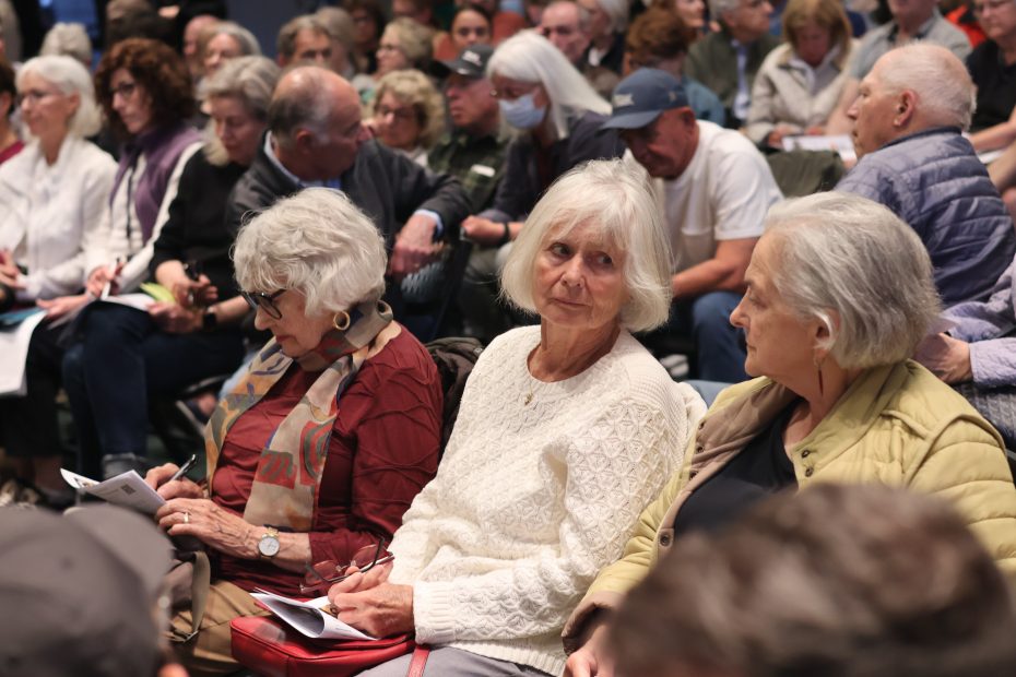 A large group of older adults seated closely together indoors, some reading papers, others engaged in conversation, at what appears to be a community meeting or event.