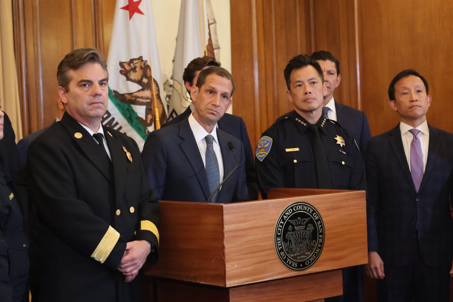 A group of officials, including fire and police chiefs, stand at a podium with the seal of San Francisco County during a press conference; California flags are in the background.
