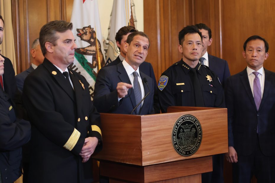 A group of officials, including uniformed personnel and a man at a podium, speak at a press conference in a room with wood-paneled walls and official seals.