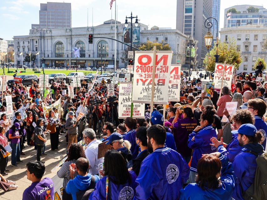 A large crowd gathers outside a city building, holding signs and banners during an immigration protest. Many participants wear matching blue jackets. Photographers and media are present.