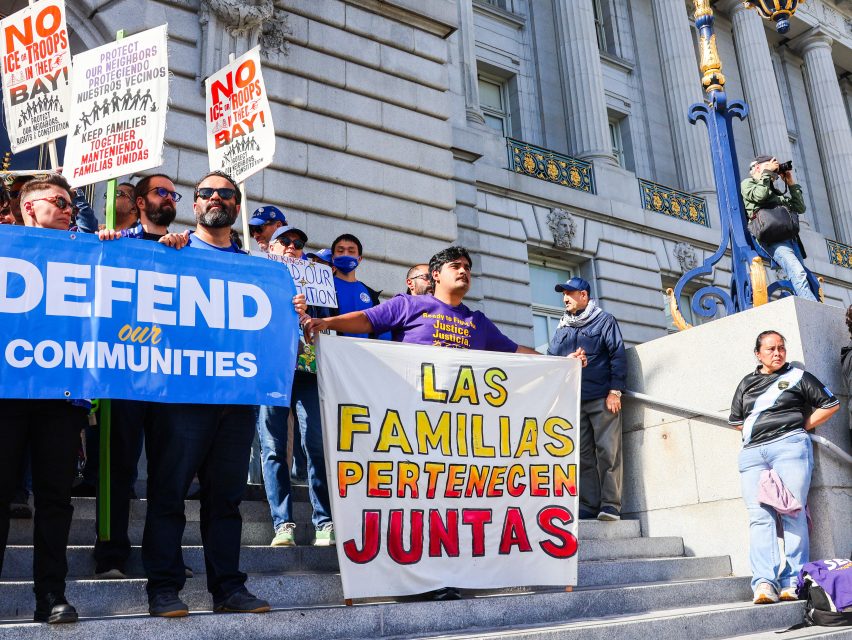 A group of people stand on steps holding signs about immigration, including one that reads “Defend Our Communities” and another in Spanish saying “Las Familias Pertenecen Juntas.”.