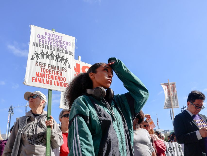 A group of people at a rally stand near signs advocating for keeping families together, protecting neighbors and communities, and supporting fair immigration policies.