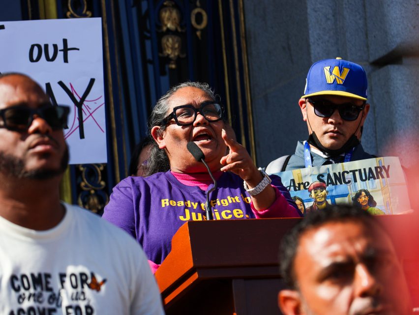 A woman speaks at a podium during a rally on immigration, wearing a purple "Ready to fight for Justice" shirt, with others and protest signs visible around her.